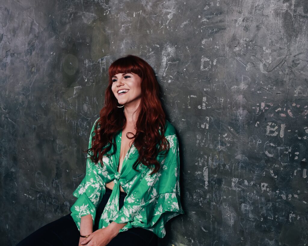 Monte in a green patterned top, smiling, leaning against a gray wall.
